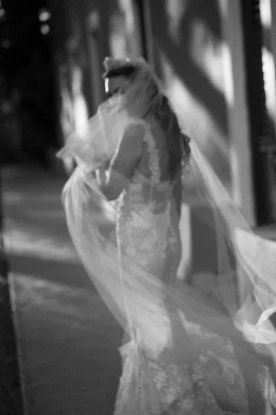 Bride portrait in a black and white bridal portrait, wearing an off-the-shoulder lace wedding dress and holding a long veil on a tree-lined walkway at night
