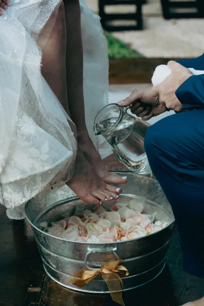Foot washing ceremony with bride’s lace dress and veil as water pours from a glass pitcher into a galvanized tub with rose petals outdoors