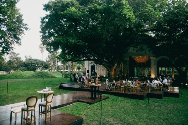 Outdoor reception setup with lounge chairs and round cocktail tables on a wood platform stage under a large tree canopy, lit by candles and tiki torches