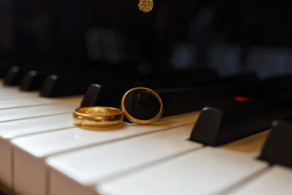 Wedding rings close-up of gold wedding bands and a groom’s signet ring resting on glossy piano keys in a music-themed detail.
