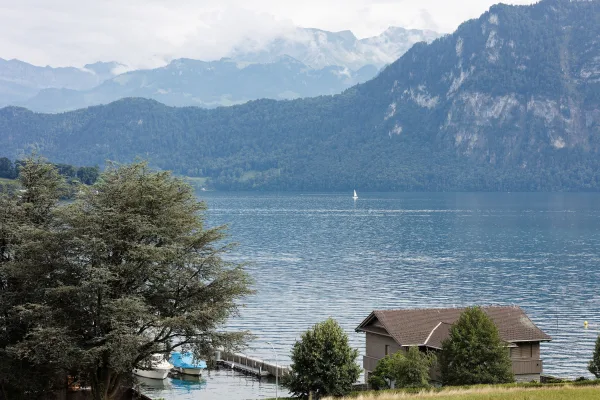 Mountain lake view with a cozy lakeside cabin, backed by forested mountains and a cloudy sky, reflected in calm blue water.