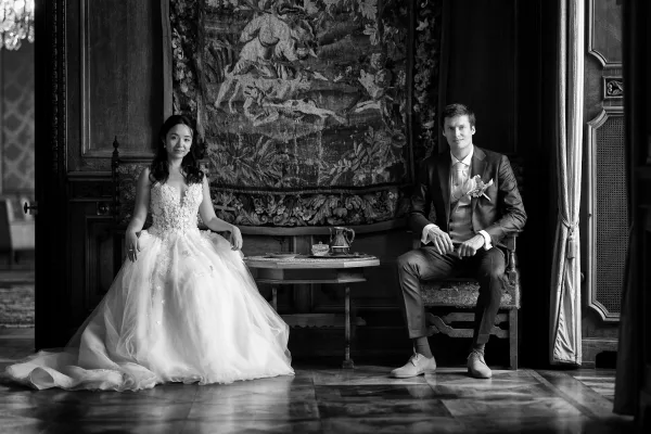 Couple portrait featuring seated bride and groom on antique chairs before a tapestry and wood-paneled backdrop, posed in elegant stillness.