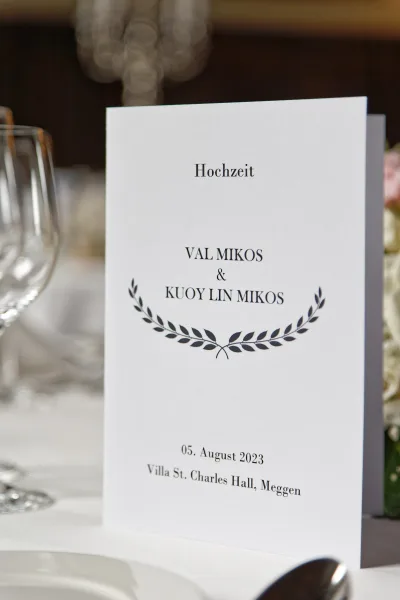 Wedding program serving as the ceremony program card at a place setting, accented by soft florals, with blurred stemware in the background.