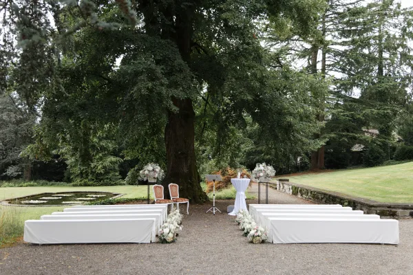 Outdoor ceremony setup beneath a grand garden tree, a ceremony under a tree with white benches facing a pond and lawn beyond.