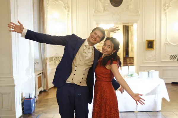 Couple portrait of a bride and groom in a fun pose, bride in a red lace dress, set against ornate white walls with sconces and a parquet floor.
