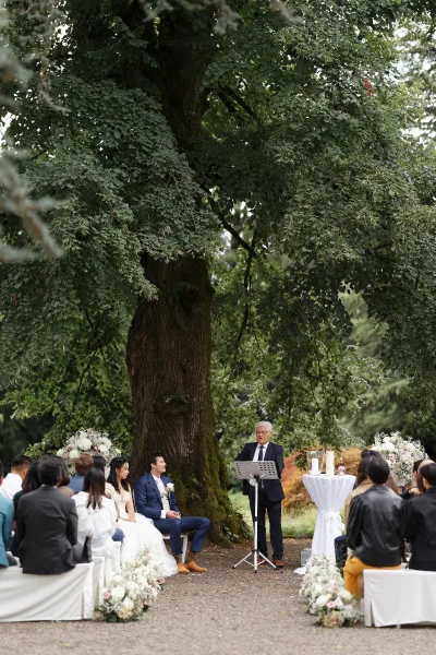 Ceremony moment at an outdoor wedding ceremony beneath a large tree, with aisle floral arrangements lining a gravel path amid lush garden greenery.