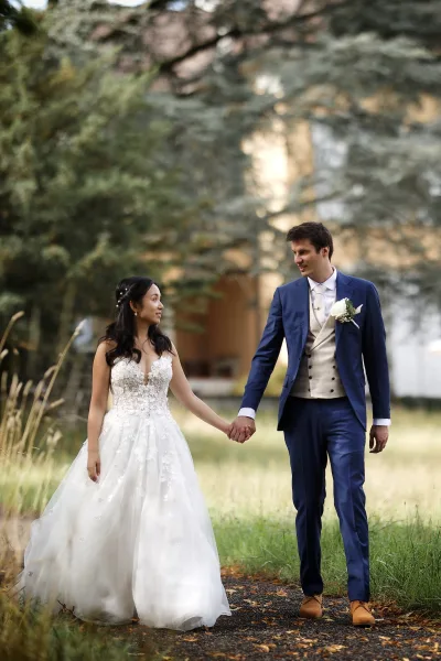 Couple portrait of the bride and groom walking hand in hand, her lace gown flowing beside him along a tree-lined path by the building exterior.