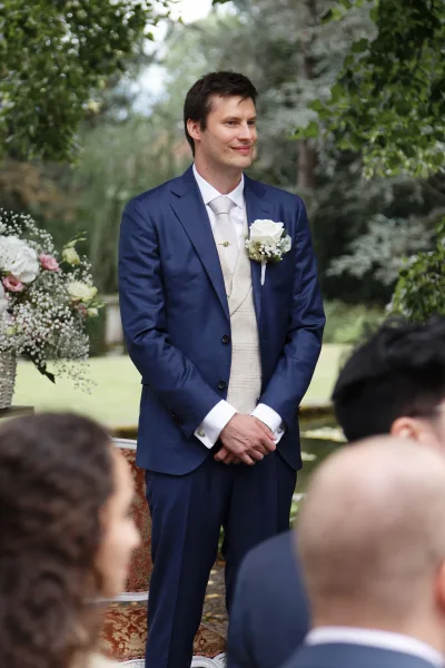 Groom portrait of a groom in a navy suit, smiling on a lawn framed by garden trees and greenery in a serene garden setting.