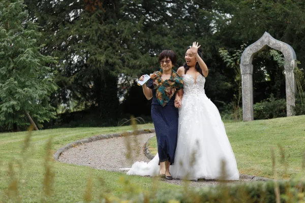 Ceremony entrance with the bride walking with mother, mother's floral scarf bright against a gravel garden path toward a stone arch.