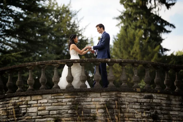 Couple portrait of a bride and groom holding hands on a stone bridge, his boutonniere vivid against the trees and sky.