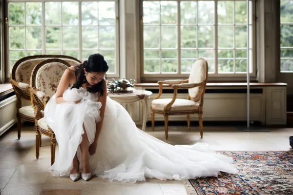 Bride getting ready, bride putting on shoes on an antique settee by large windows with garden trees and a patterned rug underfoot.
