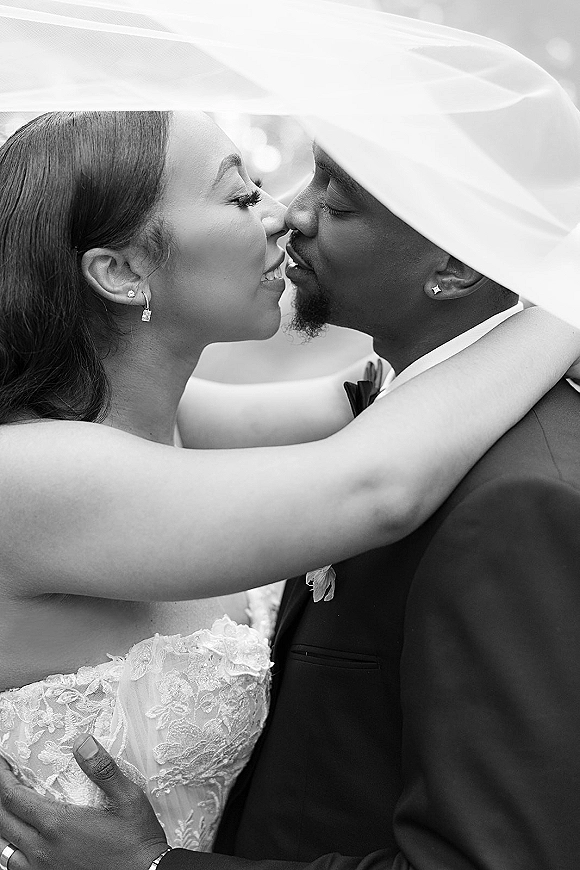 Wedding kiss portrait in black and white, bride and groom embracing under her veil, lace dress and tuxedo framed by soft bokeh lights