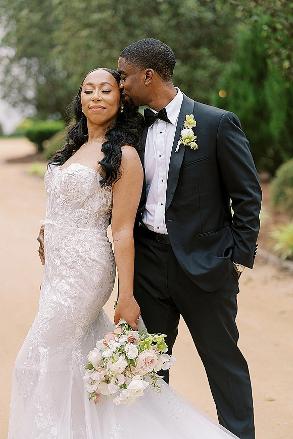 Couple portrait of groom kissing bride’s forehead as she holds a rose bouquet in a strapless lace gown on a tree-lined walkway