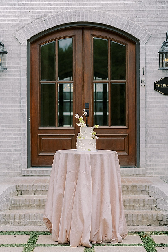 Wedding cake with white buttercream frosting, two-tier design and floral topper on a table before arched wooden doors and white brick wall