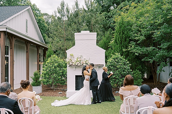 Ceremony kiss at an outdoor wedding ceremony as bride in strapless lace gown and groom in black tuxedo kiss before officiant by white brick fireplace altar