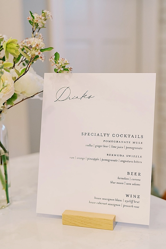 Wedding bar menu and signature drinks sign in a wooden holder on a white tablecloth with a floral arrangement in soft indoor light