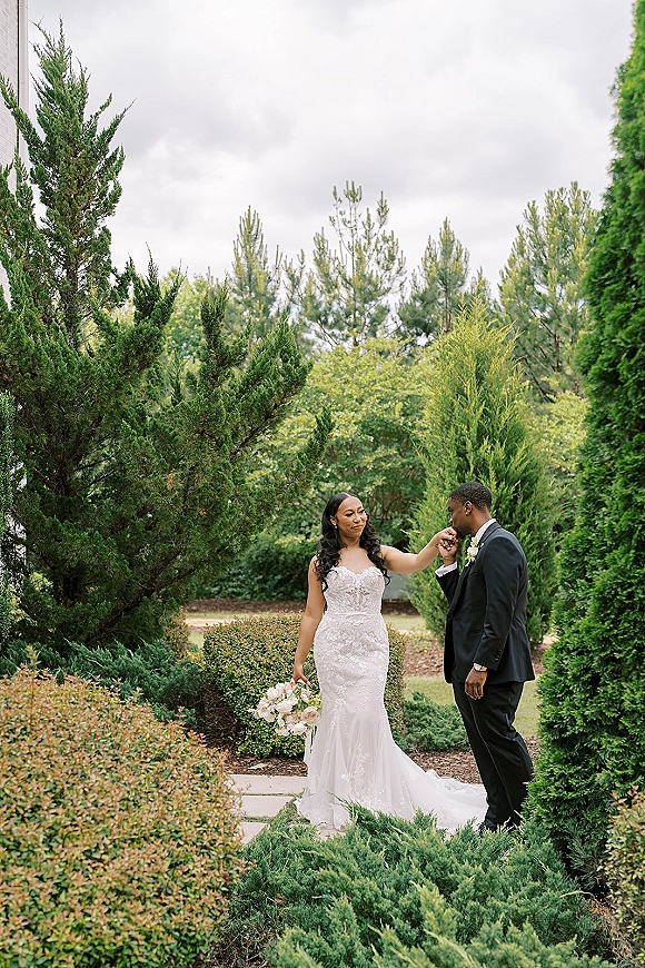 Couple portrait of bride and groom portrait as he kisses her hand, her white rose bouquet beside a strapless lace dress in a lush garden setting