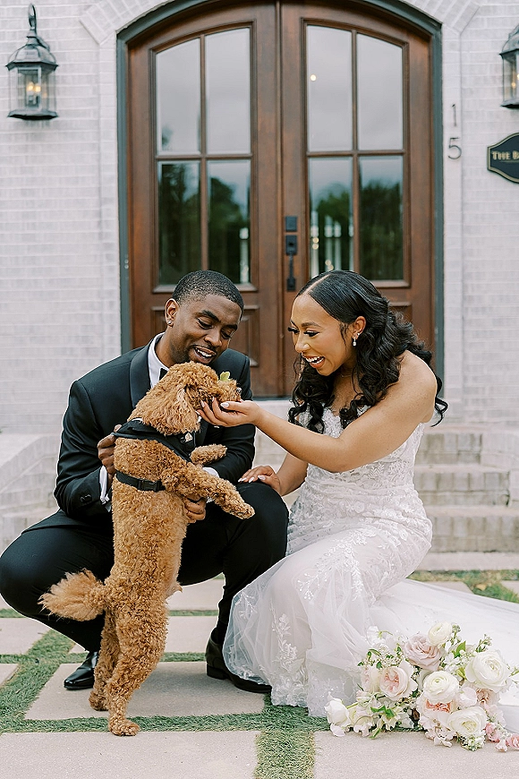 Couple portrait of bride and groom laughing as they kneel and hold their dog on brick steps by double wooden doors, bouquet in hand