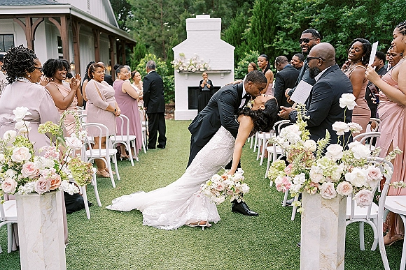 Wedding kiss as the couple shares a ceremony kiss moment in the aisle, bridal party in blush and guests cheering on a garden lawn
