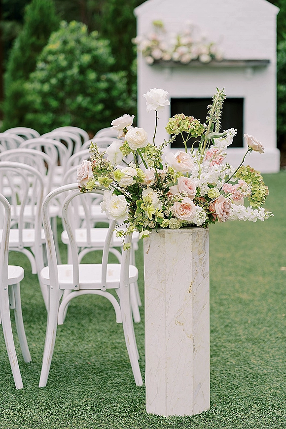 Ceremony aisle flowers in white and blush arranged on a marble pedestal, flanked by white chairs on a green lawn before a fireplace