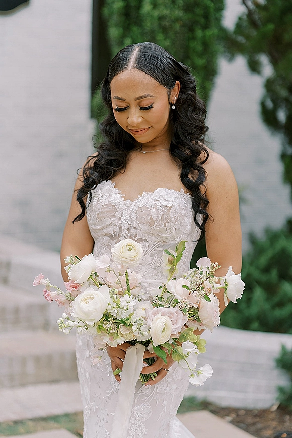 Bridal portrait of a bride holding bouquet in a strapless lace wedding dress, looking down on stone steps by a white wall and garden greenery