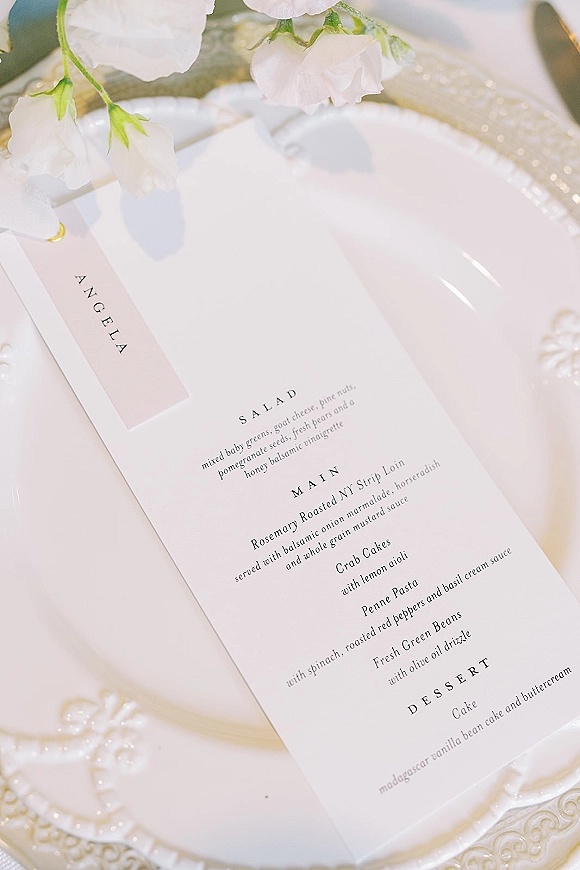 Wedding menu card with minimalist wedding menu design tied with ribbon on a gold charger, beside white plates and blush rose accents