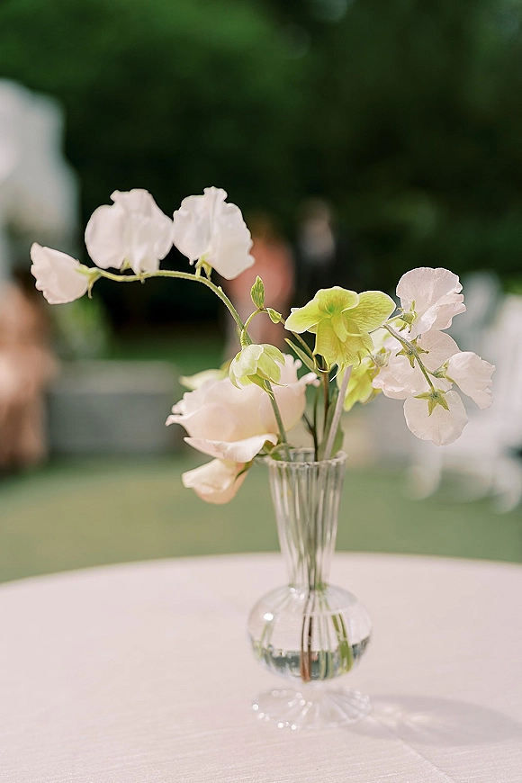 Wedding centerpiece in a clear glass bud vase with sweet pea and green hellebore blooms on a white tablecloth in a garden setting