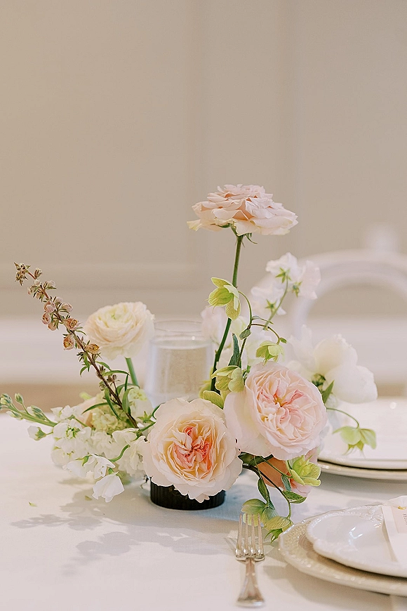 Reception centerpiece with pastel wedding centerpiece roses and greenery in a bud vase beside water glass and white place setting on tablecloth