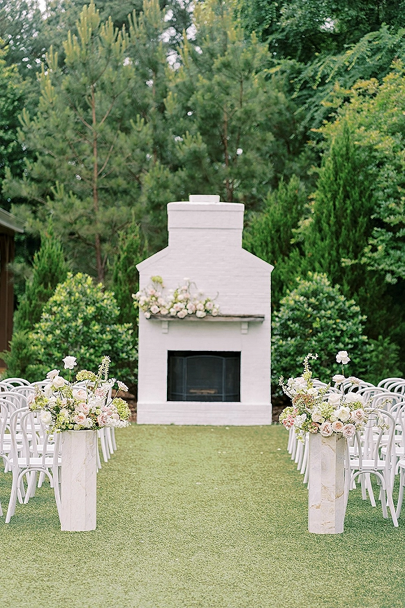 Ceremony aisle decor with white chairs and pastel rose floral arrangements on pedestal plinths, set on a garden lawn with evergreens