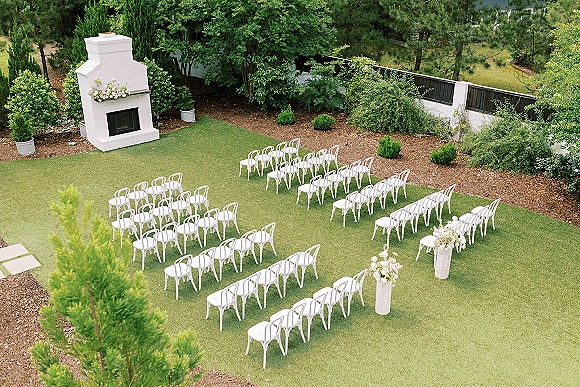 Ceremony setup for an outdoor wedding ceremony with white chairs in rows on a lawn, floral arrangements on pedestal stands by a fireplace backdrop