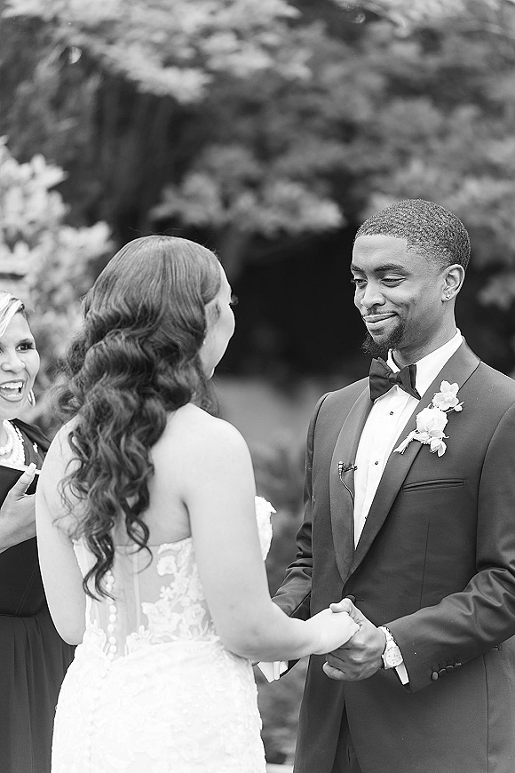 Wedding vows as bride and groom hold hands during an outdoor garden ceremony, officiant speaking into a microphone beneath trees