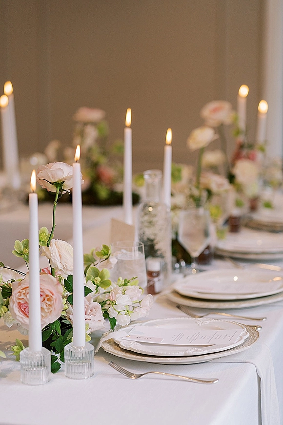 Reception tablescape with wedding table centerpiece of roses and greenery, white taper candles in glass holders, and gold-rim chargers with menus