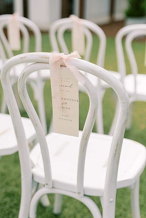 Ceremony seating sign with a reserved seat tag tied in a ribbon bow on a white wooden chair in outdoor lawn ceremony rows