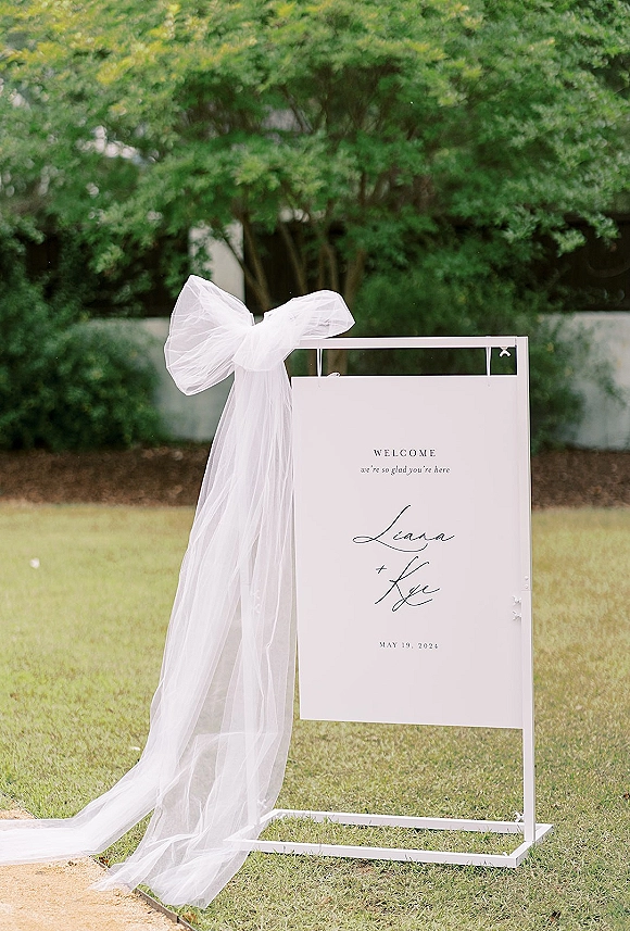 Wedding welcome sign on a white metal stand with a sheer tulle bow, set on a grassy garden lawn near greenery and a concrete wall