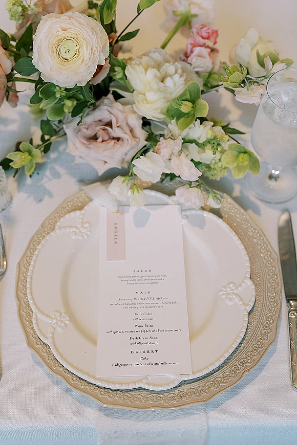 Reception tablescape with wedding place setting, menu card on scalloped plate, gold charger, silverware, goblet, and rose centerpiece