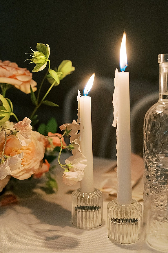 Wedding tablescape with a taper candle centerpiece, roses and sweet peas in glass holders on linen tablecloth against a dark wall