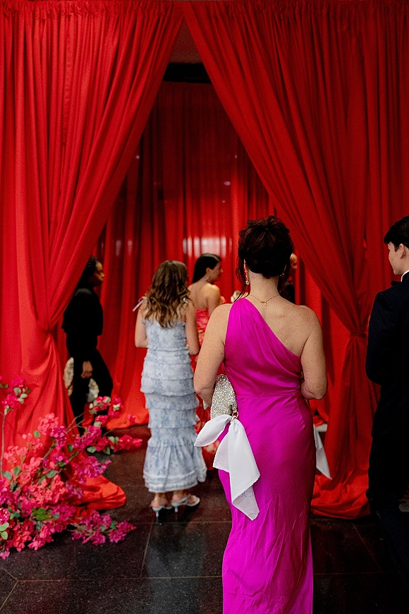 Wedding guest attire in a one shoulder wedding guest dress, holding a sparkly bow clutch with jewelry, at a red draped entrance with florals