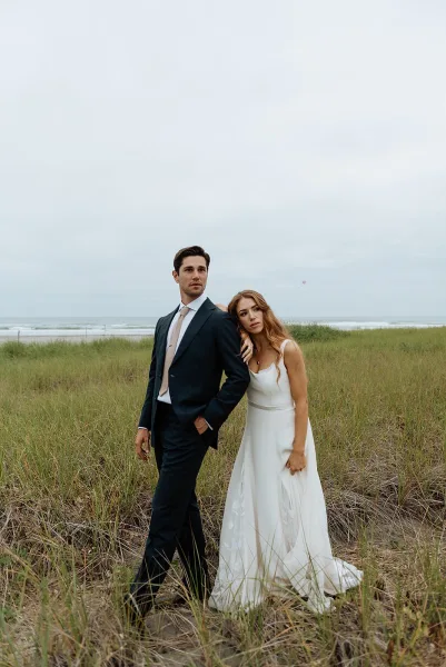 Couple portrait at a beach wedding, bride leaning on groom in a simple satin dress and black suit beside dunes and tall grass under cloudy sky