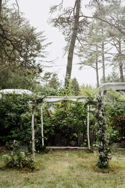 Wedding ceremony arch with greenery wedding arch garland and white draping, set on a lawn with tall trees and garden shrubs under overcast sky
