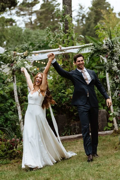 Recessional moment as bride and groom walk the aisle holding hands, bride in long veil cheering under a floral wooden arch in forest garden setting