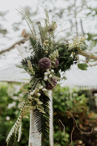 Wedding altar flowers with asymmetrical ferns and white wildflowers draped on a wooden arch, set against trees and a blurred canopy tent