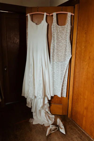 Wedding dress on hanger with sequined details and white bridal heels below, displayed in a wood wardrobe closet with tiled floor