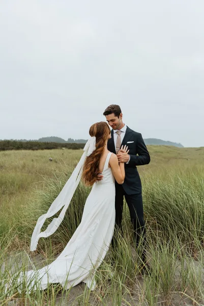 Couple portrait of bride and groom embrace on sand dunes, her long veil blowing in the wind under an overcast sky with tall grass