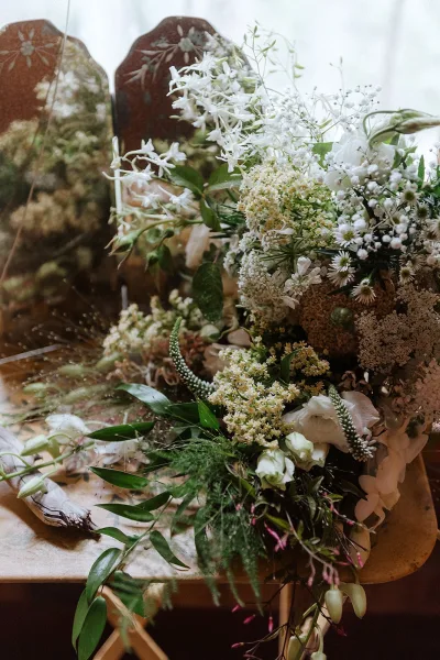 Bridal bouquet of white wildflowers with trailing greenery, arranged on a wooden tabletop beside a vintage mirror and soft outdoor greenery