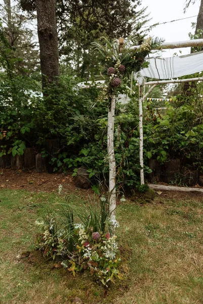 Wedding arbor decor on a birch wedding arch with asymmetrical florals and greenery, striped draping and ribbons in a woodland clearing