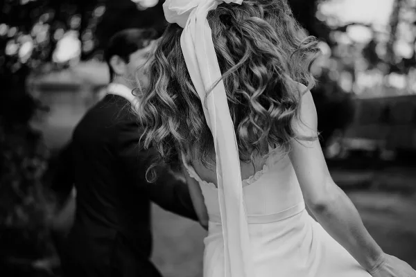 First dance moment as bride and groom dancing outdoors, bride seen from behind in strapless dress with long ribbon hair bow, trees and guests blurred