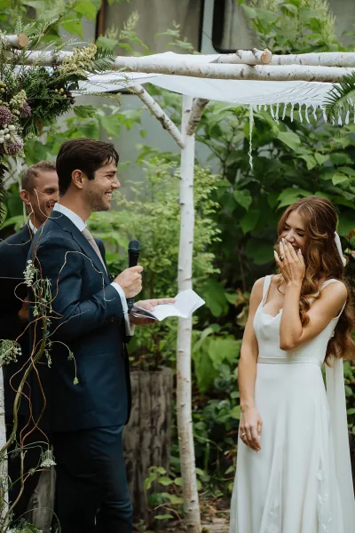 Wedding vows as the groom reading vows into a microphone beside an emotional bride under a birch chuppah with greenery outdoors
