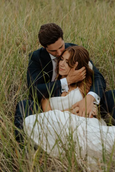 Couple portrait of bride and groom cuddle with a forehead kiss, seated in tall dried grass, her wedding dress against his dark suit