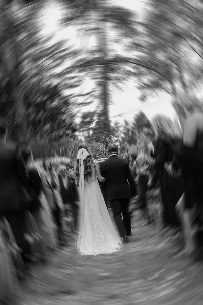 Processional moment as bride walking down aisle arm in arm, long dress train and hair ribbon beneath trees with guests seated