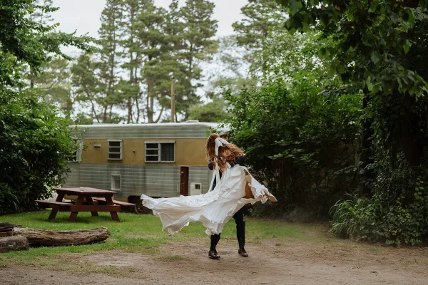 Couple portrait of the groom lifting bride as her wedding dress swirls, outdoors on a dirt path near a vintage camper in greenery